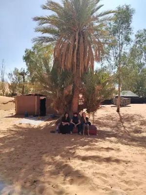 three people sitting in the sand under a palm tree at Oasis Desert Camp Obira in Merzouga