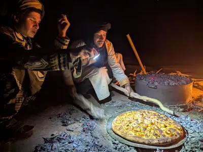 two men are cooking a pizza on a fire at Camp Local Family in Mhamid