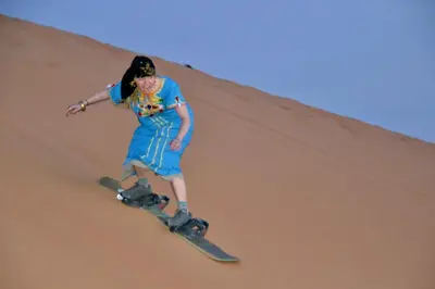 a woman riding a snowboard down a sand dune at Sahara Luxury Camp - Nuit de rêve dans le désert de Merzouga ! in Merzouga