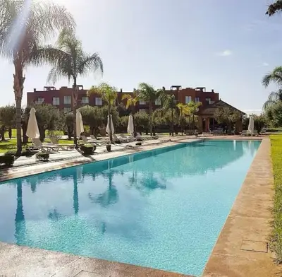 a large blue swimming pool with palm trees and umbrellas at Appartement Cosy avec grande terrasse et piscine en face du stade Can2025 in Dar Rahal Ben Saïd