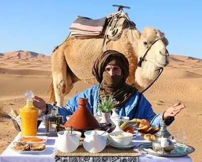 a man sitting at a table with a camel in the desert at Erg Chebbi Luxury Package & Activities in Merzouga