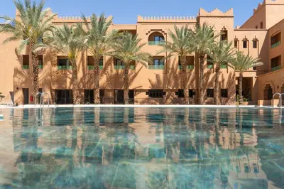 a swimming pool with palm trees in front of a building at Amanar Hôtel & Spa Ouarzazate in Ouarzazate