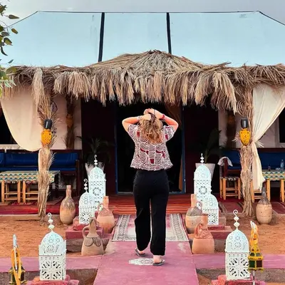 a woman is standing in front of a thatch umbrella at Sunlight Luxury Desert Camp in Merzouga