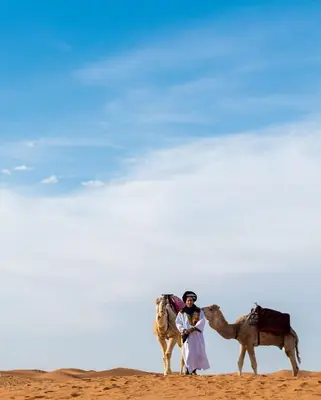 a man standing next to two camels in the desert at The Desert Palace in Merzouga