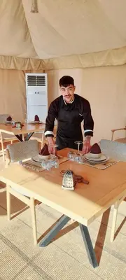 a man standing over a wooden table with plates on it at Camel desert camp in Merzouga