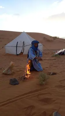 a man sitting next to a fire in front of a tent at Isolé Bivouac Merzouga in Merzouga
