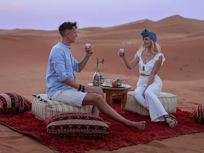 a man and woman sitting in the desert holding drinks at Global Desert Luxury Camp in Merzouga