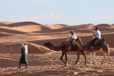 three people are riding on camels in the desert at Merzouga Luxury Desert Campsite in Merzouga