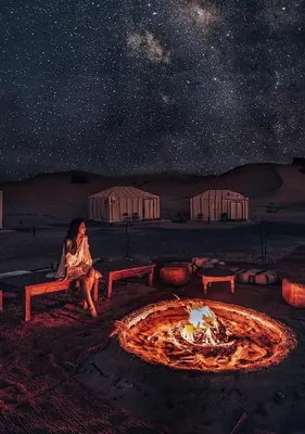 a woman sitting next to a fire pit at night at Erg Palace Luxury Camp in Merzouga