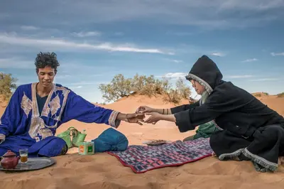 two men sitting on the sand in the desert at Desert Tours & Camp Chraika in Mhamid