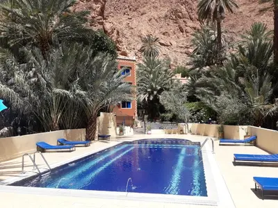 a swimming pool with palm trees and a mountain at RIAD Camping ATLAS in Tinerhir