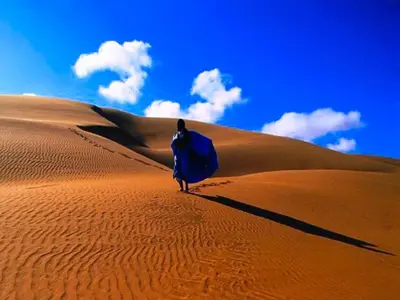 a person walking on a sand dune with a surfboard at Camp Iguidi Tours in Mhamid
