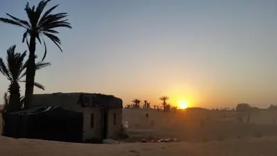 a sunset in the desert with a building and a palm tree at SaharaTime Camp in Hassilabied