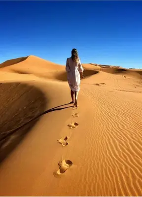 a woman walking through the desert with footprints in the sand at MorokoTour Camp in Merzouga