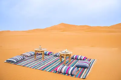 a blanket in the middle of the desert with two tables at Aiour Luxury Camp in Merzouga