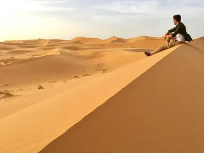 a man sitting on a sand dune in the desert at Visit Morocco Sahara Camp in Merzouga