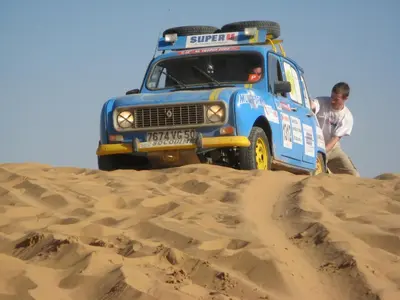 a man standing next to a blue truck in the sand at La Vallée des Dunes - Auberge, randonnées et circuits in Merzouga