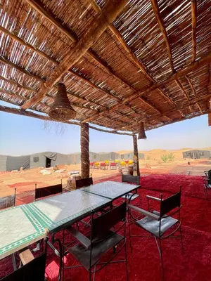 a table and chairs under a wooden roof in the desert at Mhamid Sahara Luxury Camp - Chez Naji in Mhamid