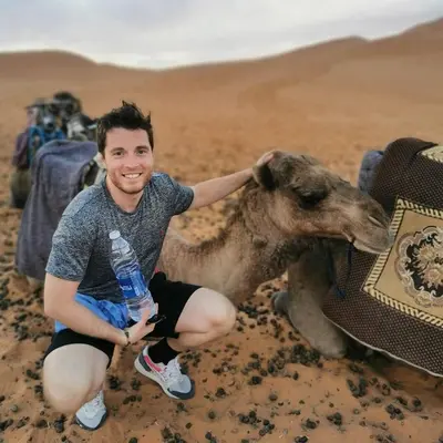 a man holding a water bottle while kneeling next to a camel at Chigaga Desert Camp in Mhamid