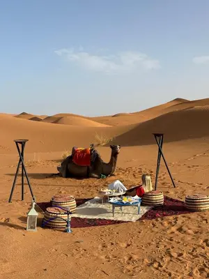 a child sitting on a blanket in the desert with a camel at Original Desert Camp in Merzouga
