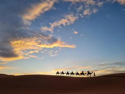 a group of people riding camels in the desert at sunset at Nomada Desert Camp in Merzouga