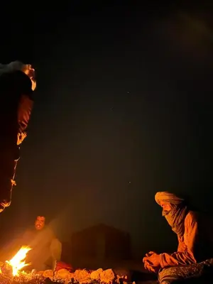 a person sitting next to a fire at night at Mhamid Sahara Camp - Mhamid El Ghizlane in Mhamid