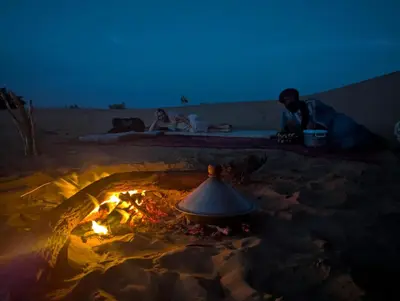 a man sitting next to a fire in the sand at Mhamid Sahara Camp - Mhamid El Ghizlane in Mhamid