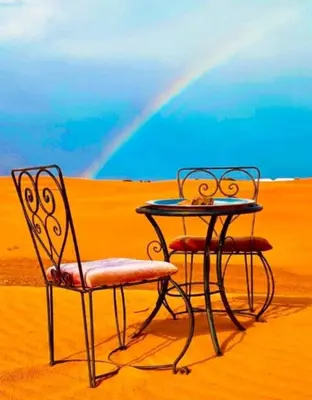 a table and chair in the desert with a rainbow in the background at Merzouga Sahara Camp in Merzouga